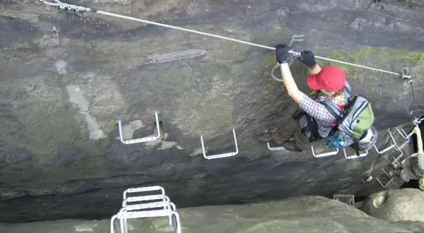 Ein Klettersteig im Elbsandsteingebirge: Eine Person sichert sich an einem Stahlseil, während sie über Felsstufen klettert.