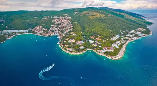 Blick auf eine Küstenstadt mit roten Dächern und grünen Hügeln, umgeben von türkisfarbenem Wasser. Ein Boot hinterlässt eine Spur im Meer. Ein echter **Istrien Geheimtipp**.