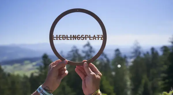 Hände halten ein Schild mit "LIEBLINGSPLATZ" vor einer malerischen Bergkulisse. Ein echter Geheimtipp im Bayerischen Wald.