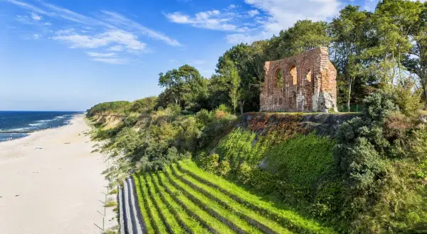 Ruinen einer alten Kirche thronen über einem Strand an der polnischen Ostsee. Terrassierte grüne Hänge führen zum Meer.