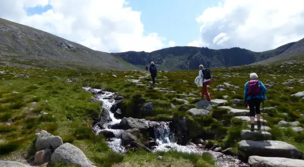 Drei Wanderer genießen das Cairngorms National Park Wandern entlang eines steinigen Pfades mit einem plätschernden Bach.