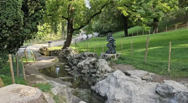 Ein kleiner Bach schlängelt sich durch den Parc des Buttes Chaumont, vorbei an Felsen und einer Skulptur. Grüne Hänge und Bäume säumen den Weg.