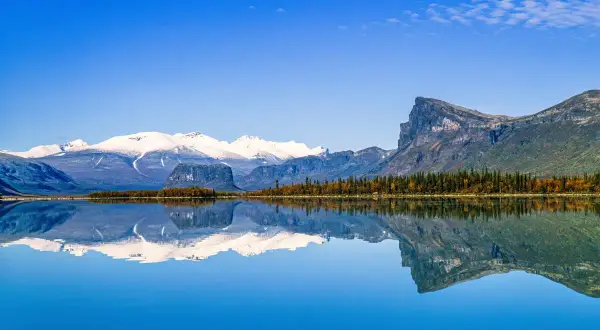 Spiegelglatte Wasserfläche reflektiert schneebedeckte Berge und herbstliche Bäume in einem schwedischen Nationalpark.