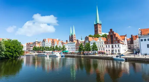 Historische Gebäude und Kirchtürme säumen das Wasser in Lübeck, einem der schönsten Ostsee Orte. Boote liegen am Kai, die Sonne scheint.
