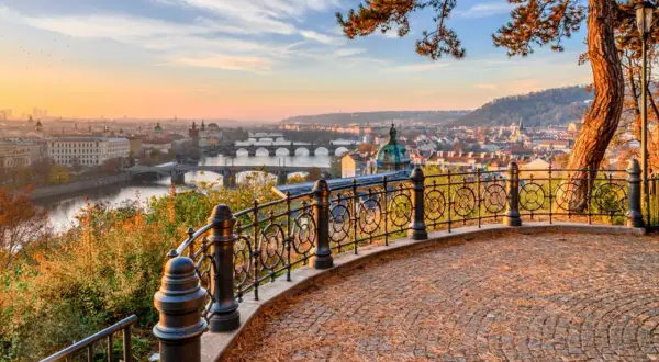 Prag im Herbst: Blick über die Moldau mit Brücken und Stadtkulisse bei Sonnenuntergang. Ein malerischer Ausblick von einem Aussichtspunkt mit gepflastertem Boden und schmiedeeisernem Geländer.