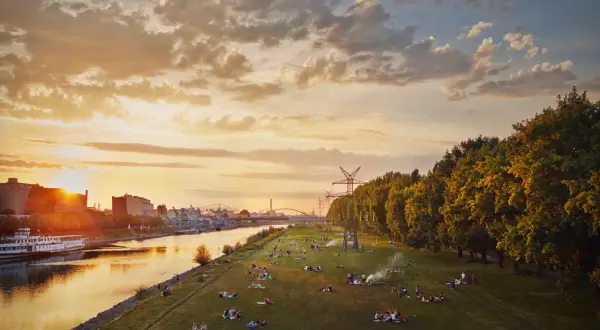 Sonnenuntergang an der Neckarwiese Mannheim: Menschen entspannen auf der Wiese, ein Schiff fährt auf dem Fluss, im Hintergrund Industrie und Bäume.