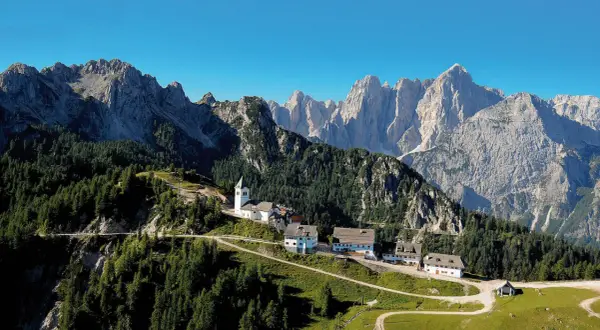 Alpe-Adria-Trail Erfahrungsbericht: Malerisches Bergdorf mit Kirche und Häusern, umgeben von Nadelwäldern und imposanten Gipfeln.