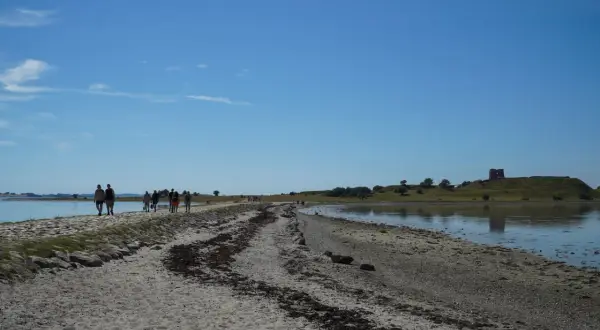 Menschen spazieren auf einem sandigen Weg entlang des Wassers in Richtung der Mols Bjerge mit einer alten Burgruine auf dem Hügel.