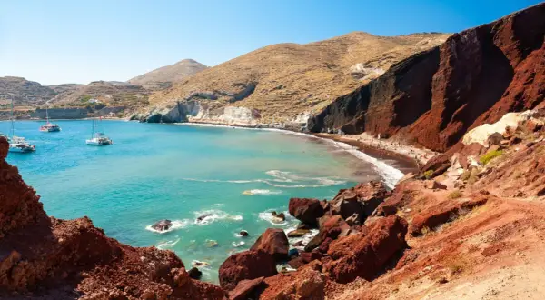 Der Rote Strand von Santorini mit türkisfarbenem Wasser, Segelbooten und Menschen, die sich auf dem roten Sand entspannen.