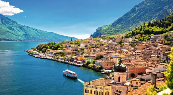 Buntes Dorf am Seeufer, Berge im Hintergrund. Ein Schiff fährt auf dem Wasser. Ein malerischer Ort am Lago Maggiore oder Gardasee.