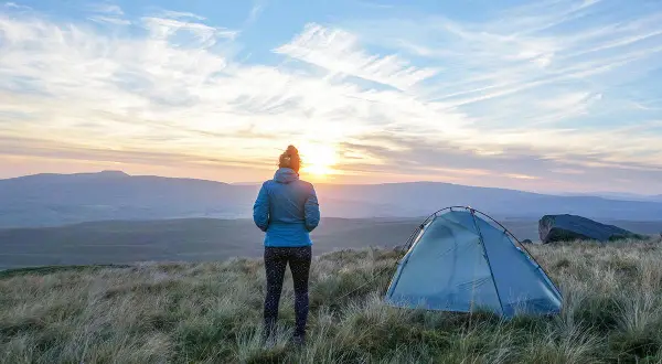 Frau blickt auf Sonnenuntergang, ihr Zelt steht bereit für das Wandern mit Zelt. Die Landschaft ist hügelig, der Himmel dramatisch.
