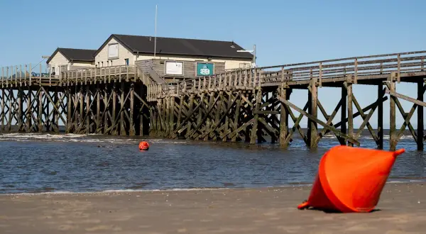 St. Peter-Ording an der Nordsee: Holzsteg mit Häusern, Bojen im Wasser und eine orangefarbene Boje am Strand.
