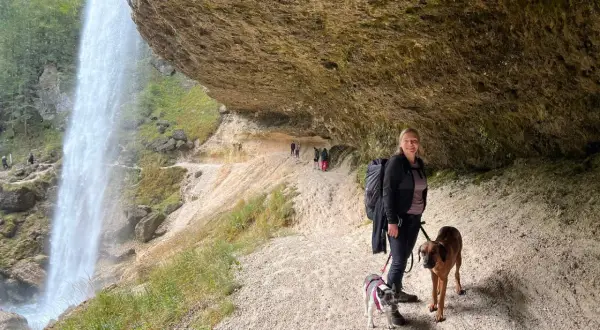 Frau mit zwei Hunden unter einem Felsvorsprung, während ein Wasserfall in der Nähe herabstürzt. Ein perfekter Ort, um die Natur zu genießen.
