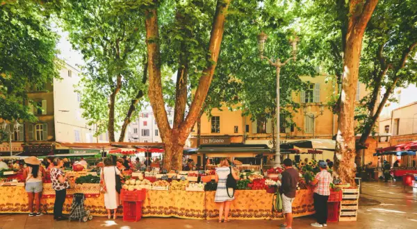 Bunter Markt in Aix-en-Provence unter schattigen Bäumen. Menschen kaufen frisches Obst und Gemüse an Ständen mit provenzalischen Stoffen.