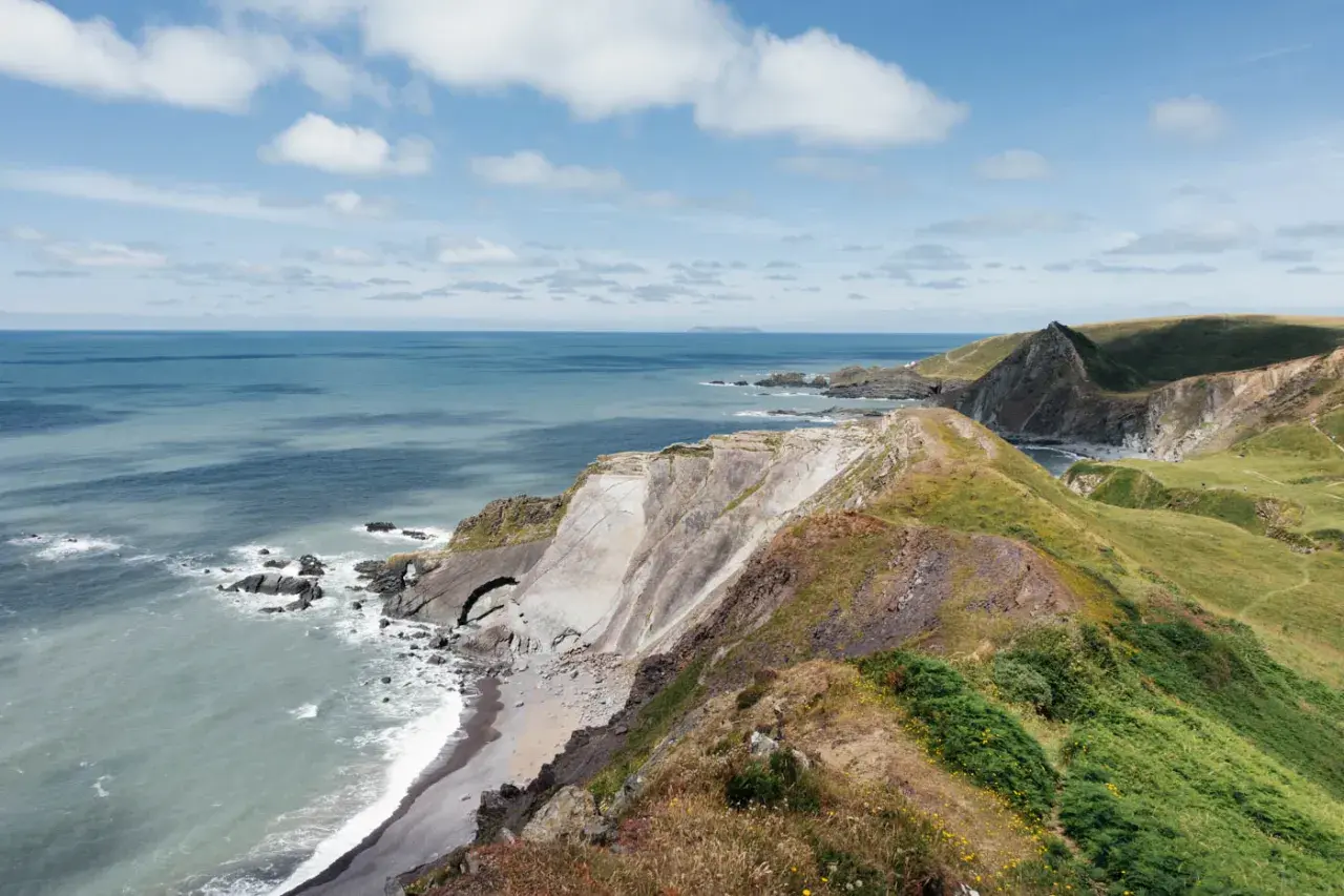 Grüne Hügel und steile Klippen am South West Coast Path. Das Meer glitzert in der Sonne, Wolken ziehen über den blauen Himmel.