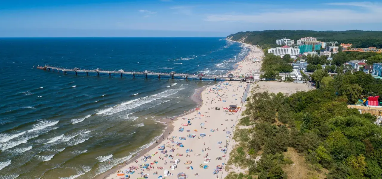 Blick auf die belebte Misdroy Promenade mit Strand, Meer und Seebrücke. Viele Menschen genießen den sonnigen Tag am Ostseestrand.