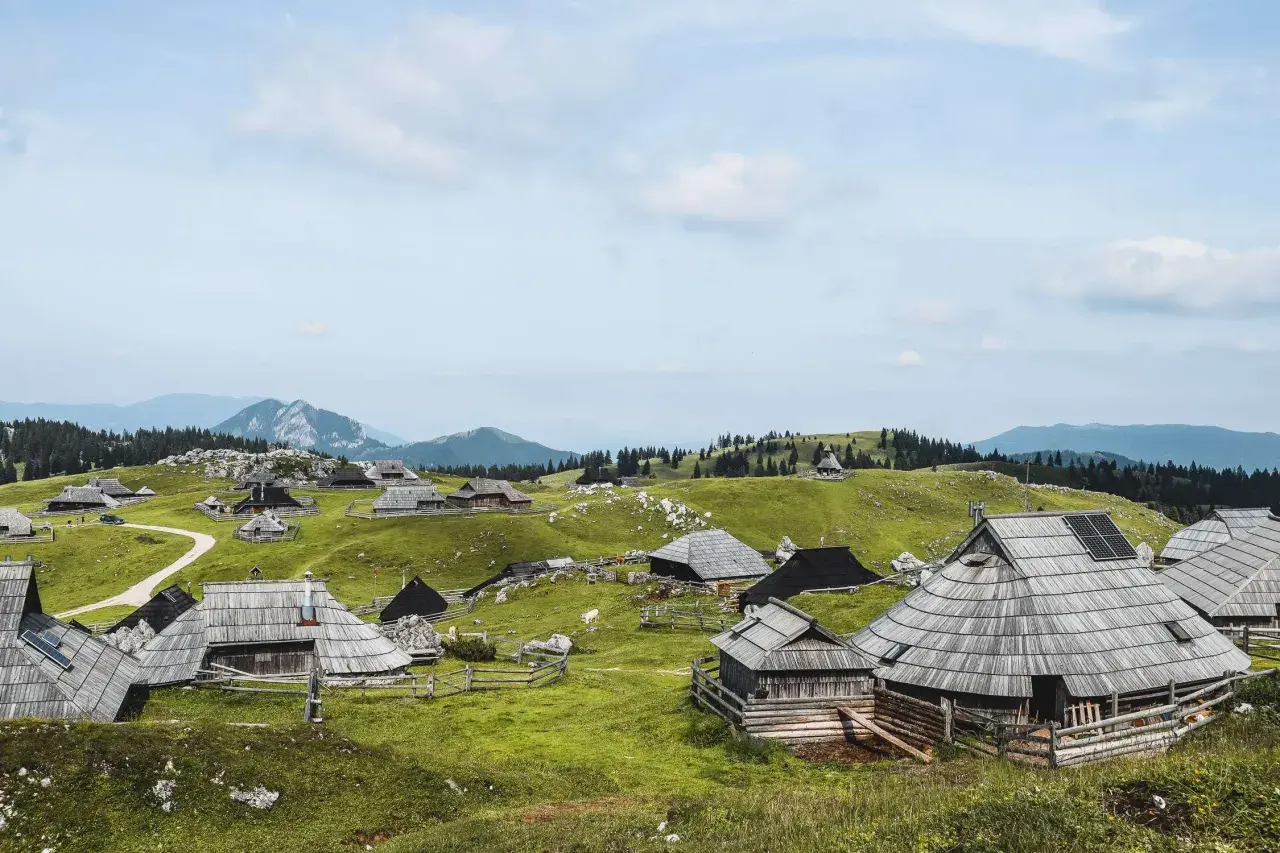 Idyllische Holzhütten auf grünen Hügeln, ein malerischer Ort für eine Velika Planina Wanderung. Berge im Hintergrund.
