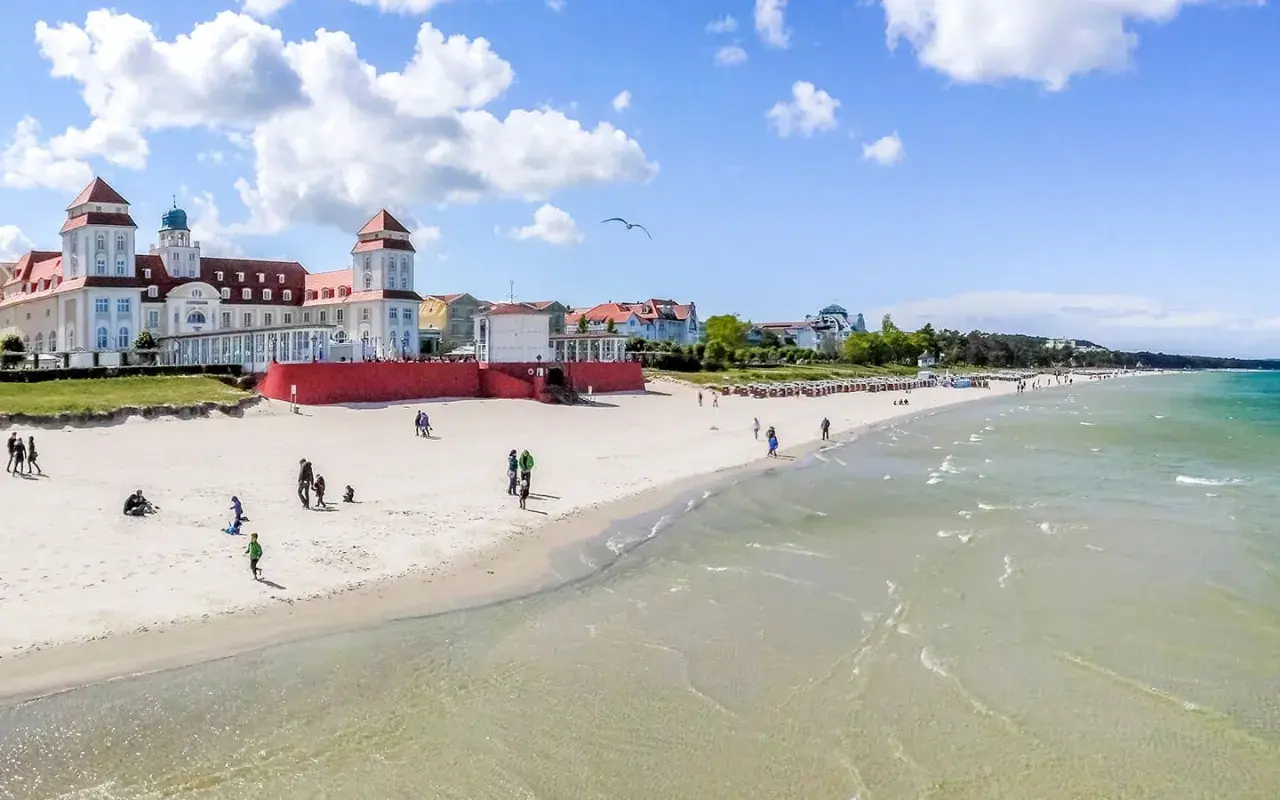 Ein sonniger Ostsee Strand mit Menschen, die spazieren gehen und Kinder spielen. Im Hintergrund ein prächtiges Gebäude im Bäderstil.