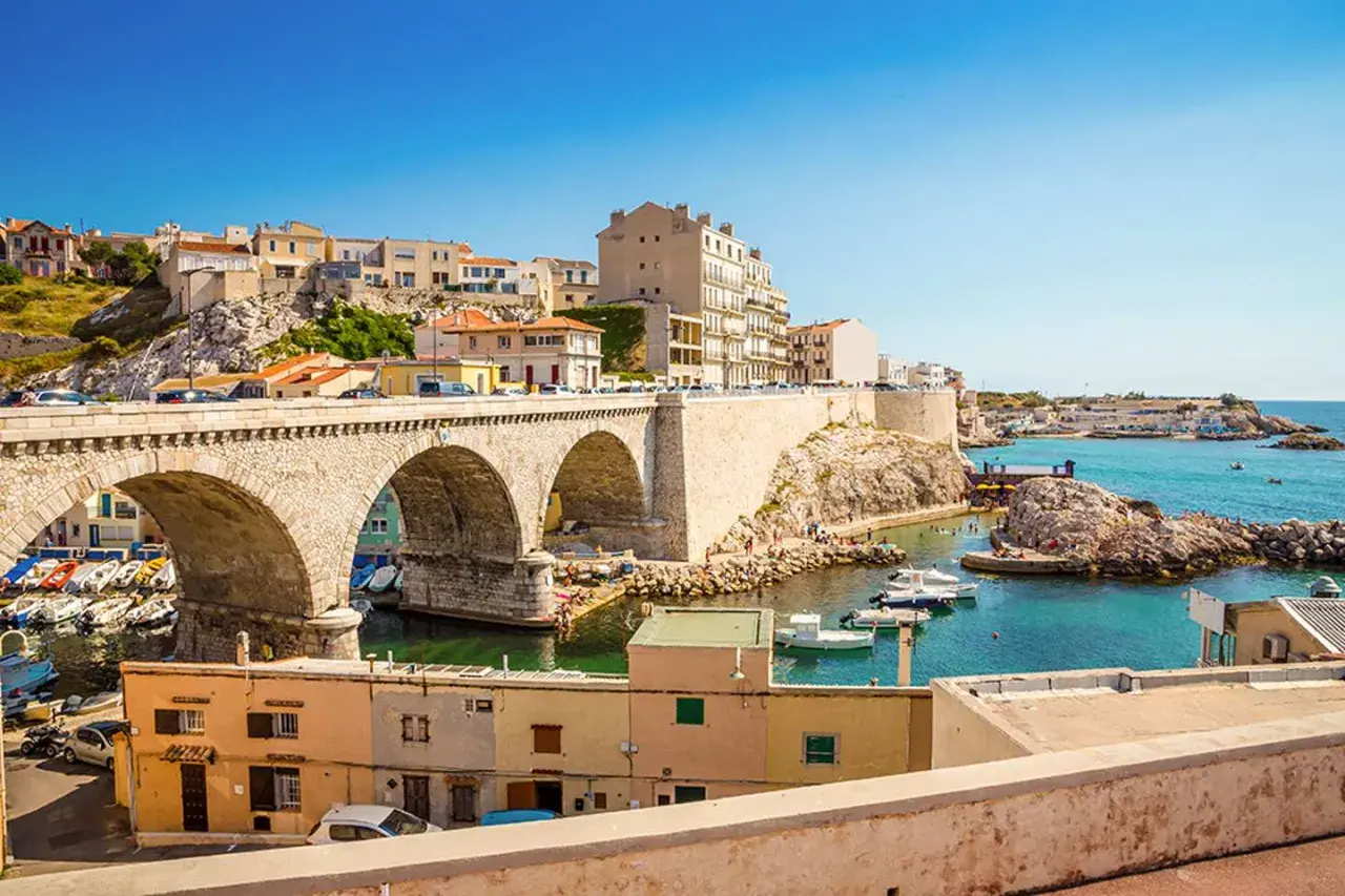 Bogenbrücke über einem Hafen mit Booten und Häusern, die sich an einen Hügel schmiegen. Die malerische marseille altstadt erstreckt sich bis zum türkisfarbenen Meer.