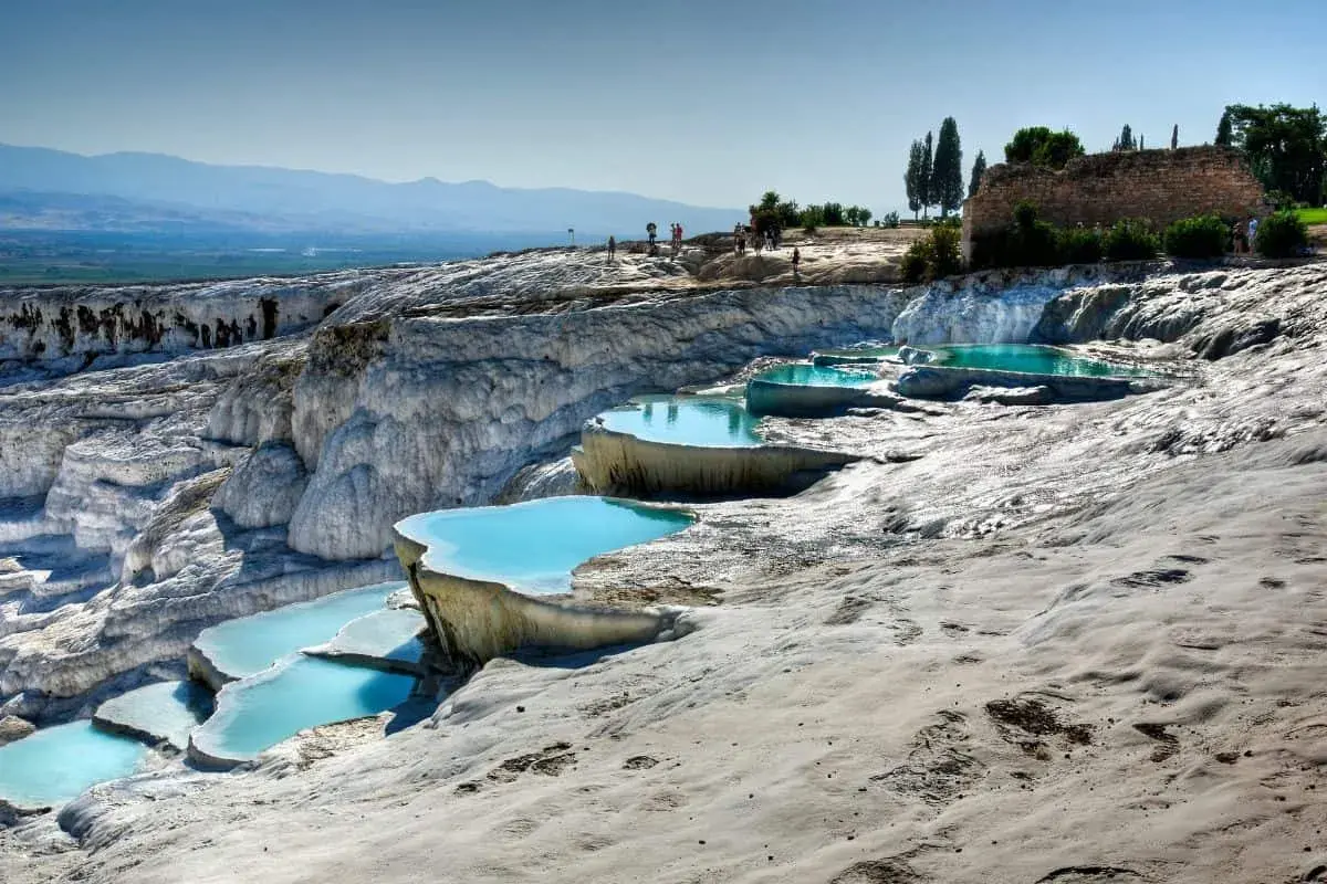 Travertinterrassen von Pamukkale heute: Türkisblaue Pools auf weißen Kalksteinterrassen, mit Besuchern im Hintergrund und antiken Ruinen.