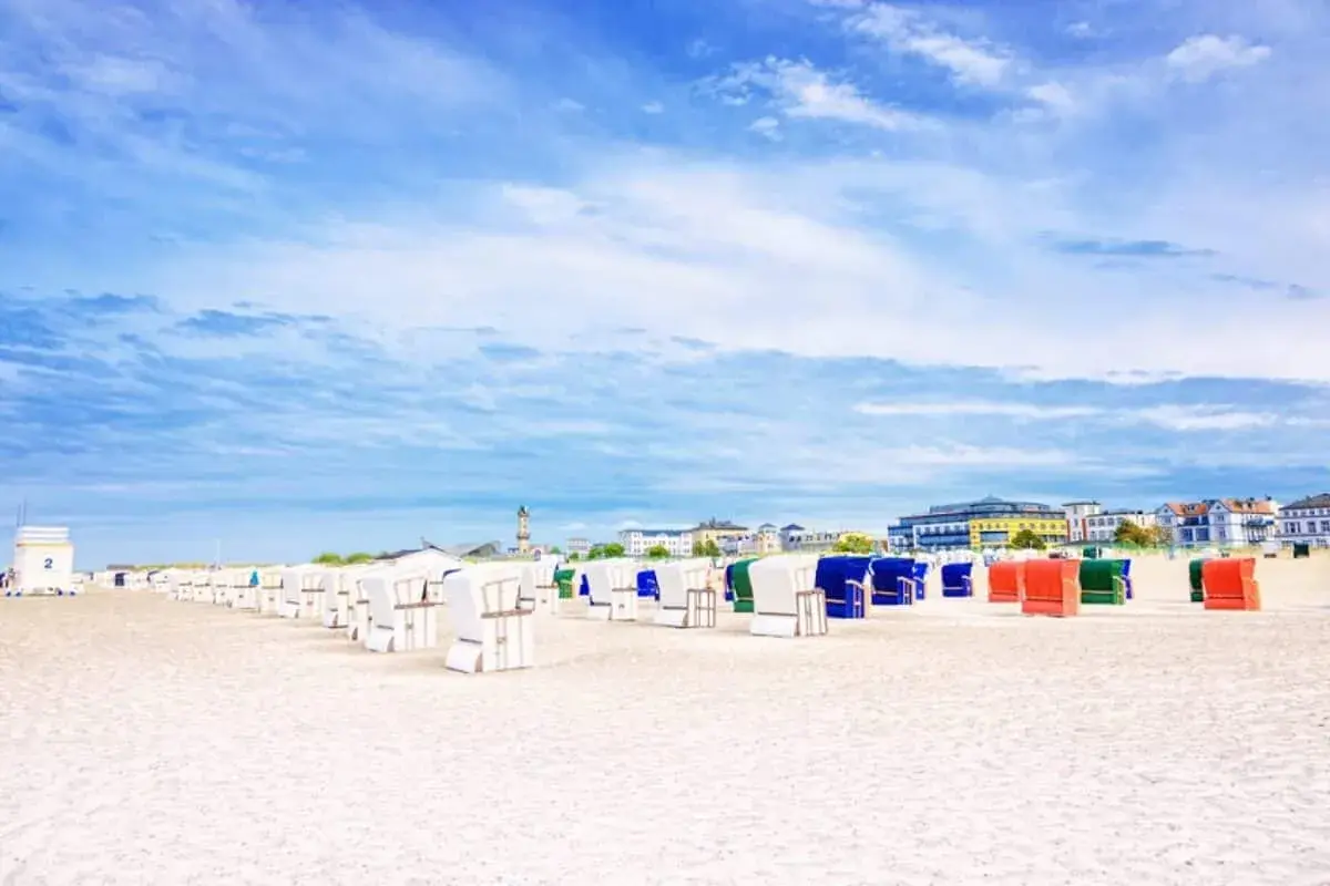Strandkörbe in bunten Farben säumen den Sandstrand eines Ostseebads in Mecklenburg-Vorpommern. Im Hintergrund die Promenade mit Häusern und ein Leuchtturm.