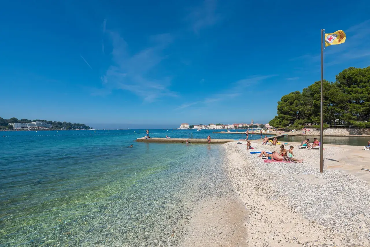 Sonniger istrien strand mit klarem Wasser, Kiesstrand und Badegästen. Im Hintergrund eine Stadtkulisse und grüne Bäume.