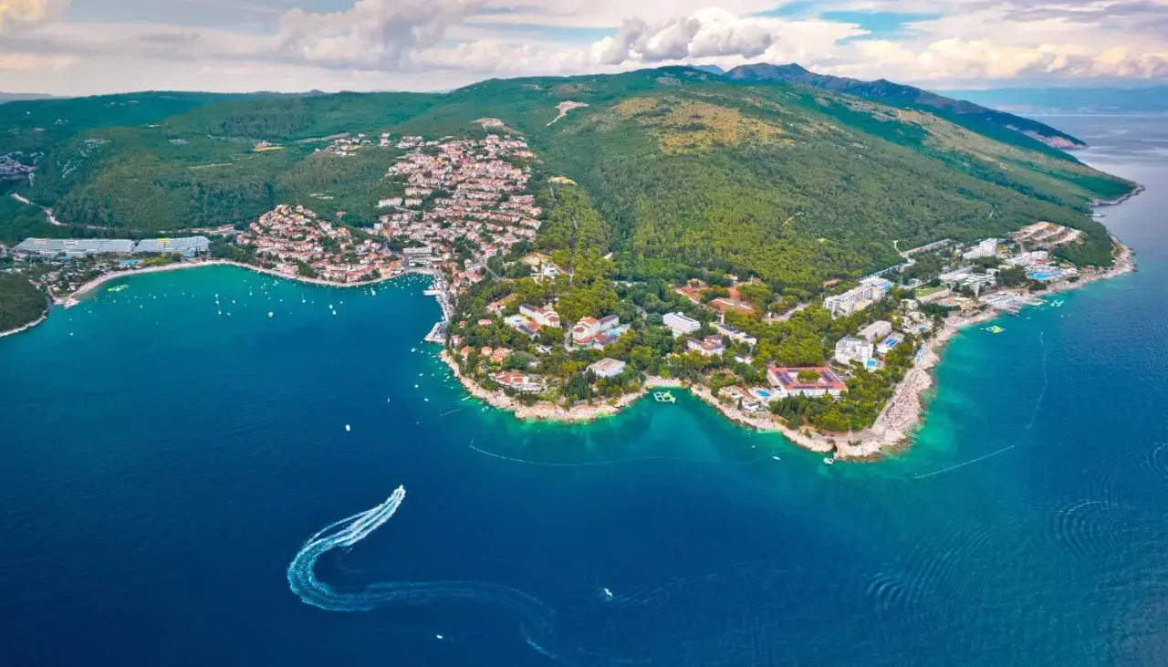 Blick auf eine Küstenstadt mit roten Dächern und grünen Hügeln, umgeben von türkisfarbenem Wasser. Ein Boot hinterlässt eine Spur im Meer. Ein echter **Istrien Geheimtipp**.