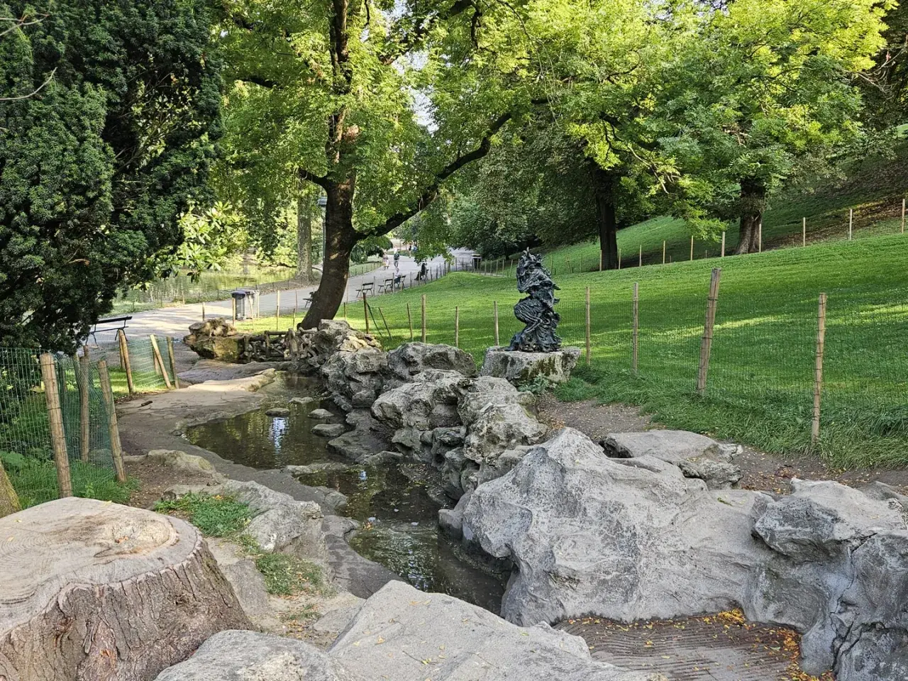 Ein kleiner Bach schlängelt sich durch den Parc des Buttes Chaumont, vorbei an Felsen und einer Skulptur. Grüne Hänge und Bäume säumen den Weg.