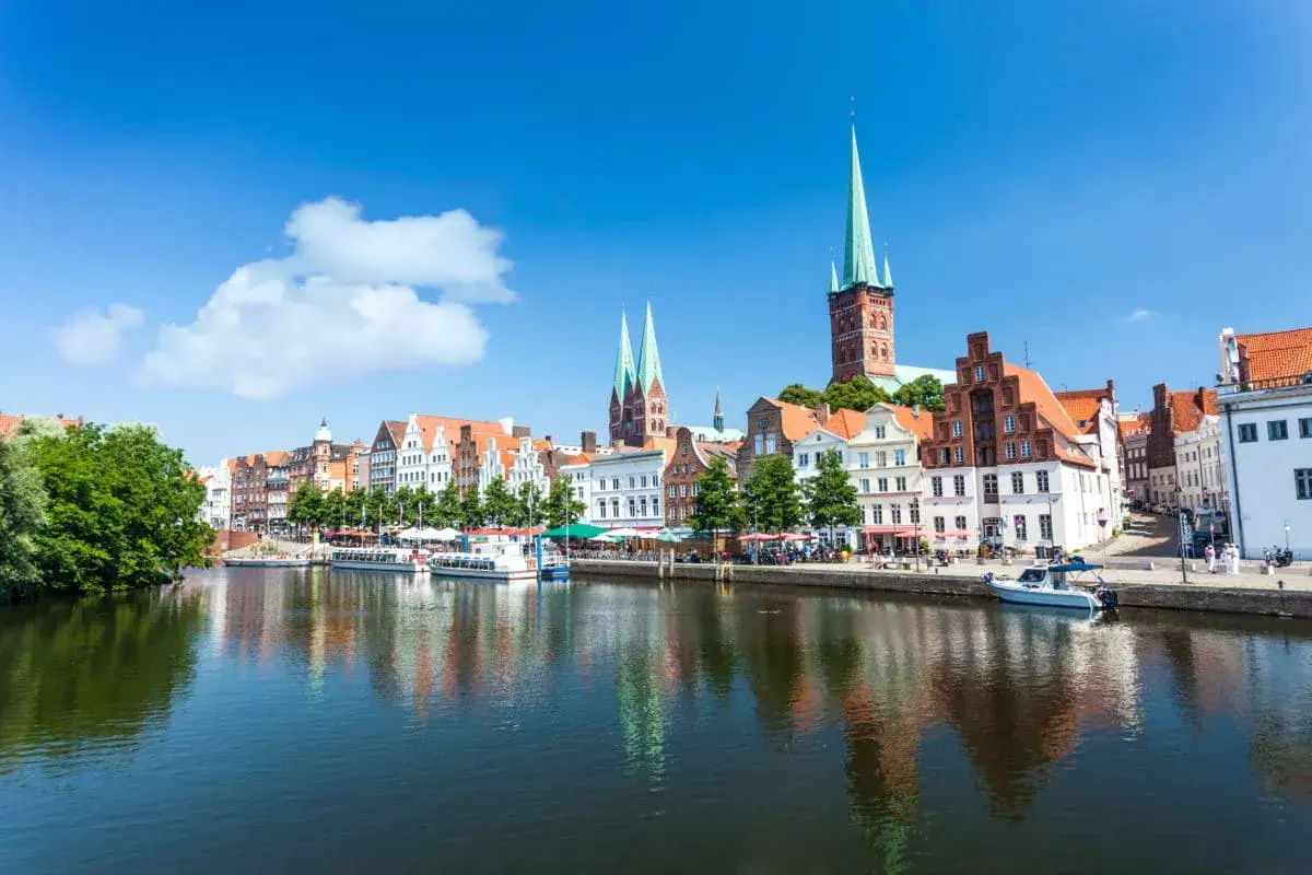 Historische Gebäude und Kirchtürme säumen das Wasser in Lübeck, einem der schönsten Ostsee Orte. Boote liegen am Kai, die Sonne scheint.
