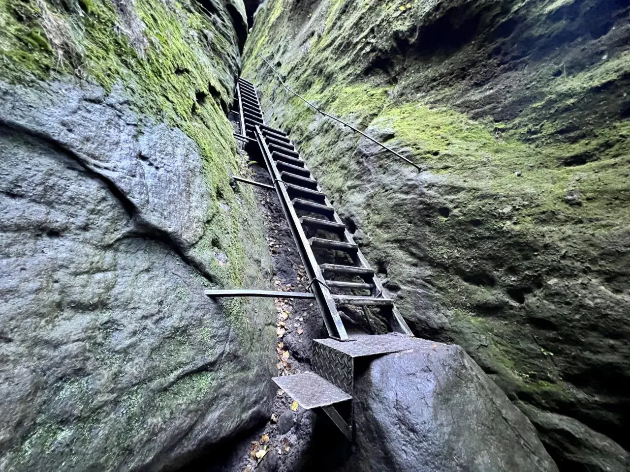 Steile Metalltreppe führt durch eine enge Felsspalte, Moos bedeckt die Wände. Eine leichte Schrammsteine Wanderung mit Ausblick.