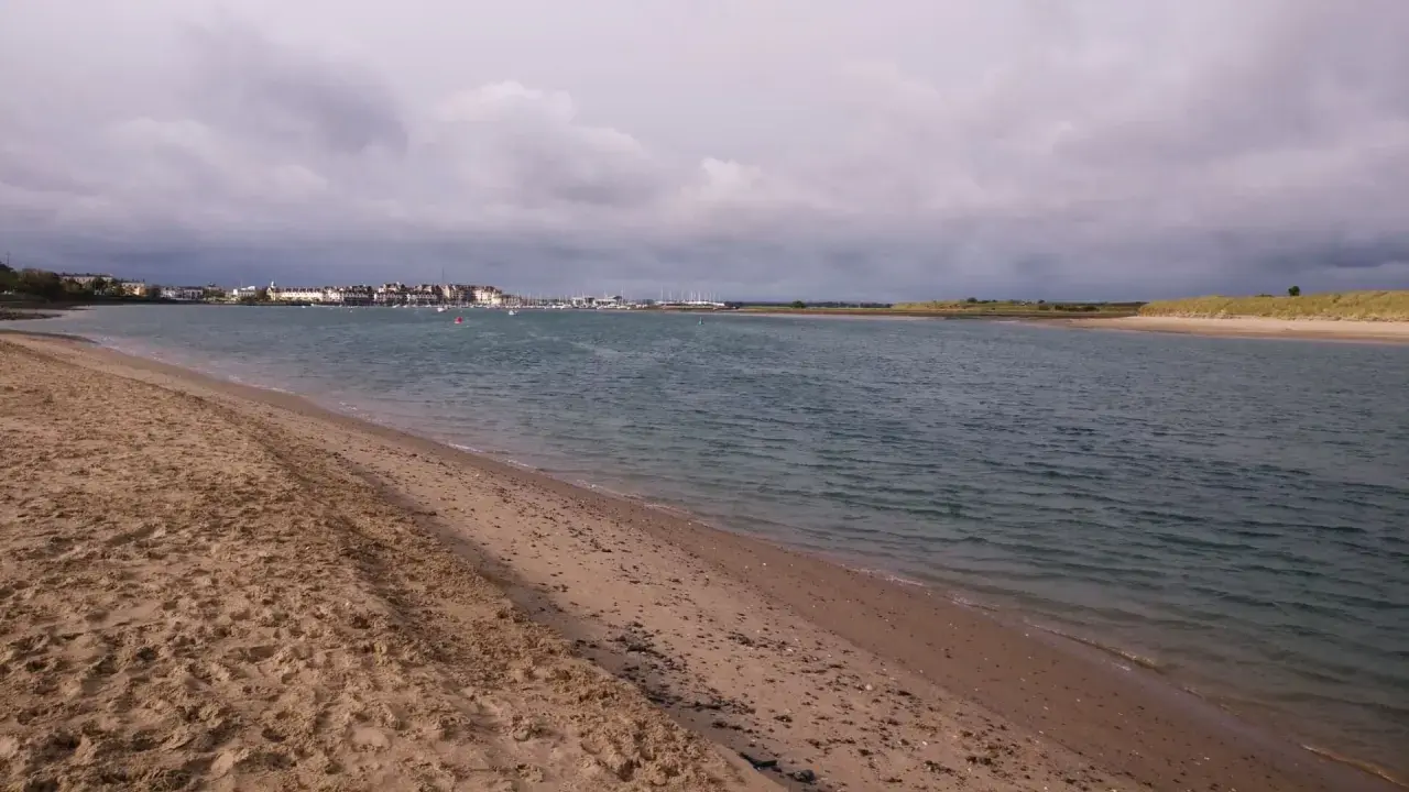 Sandiger Strand mit Blick auf eine Stadt und einen Yachthafen. Der Himmel ist bewölkt, aber das Wasser am Emden Strand ist ruhig.