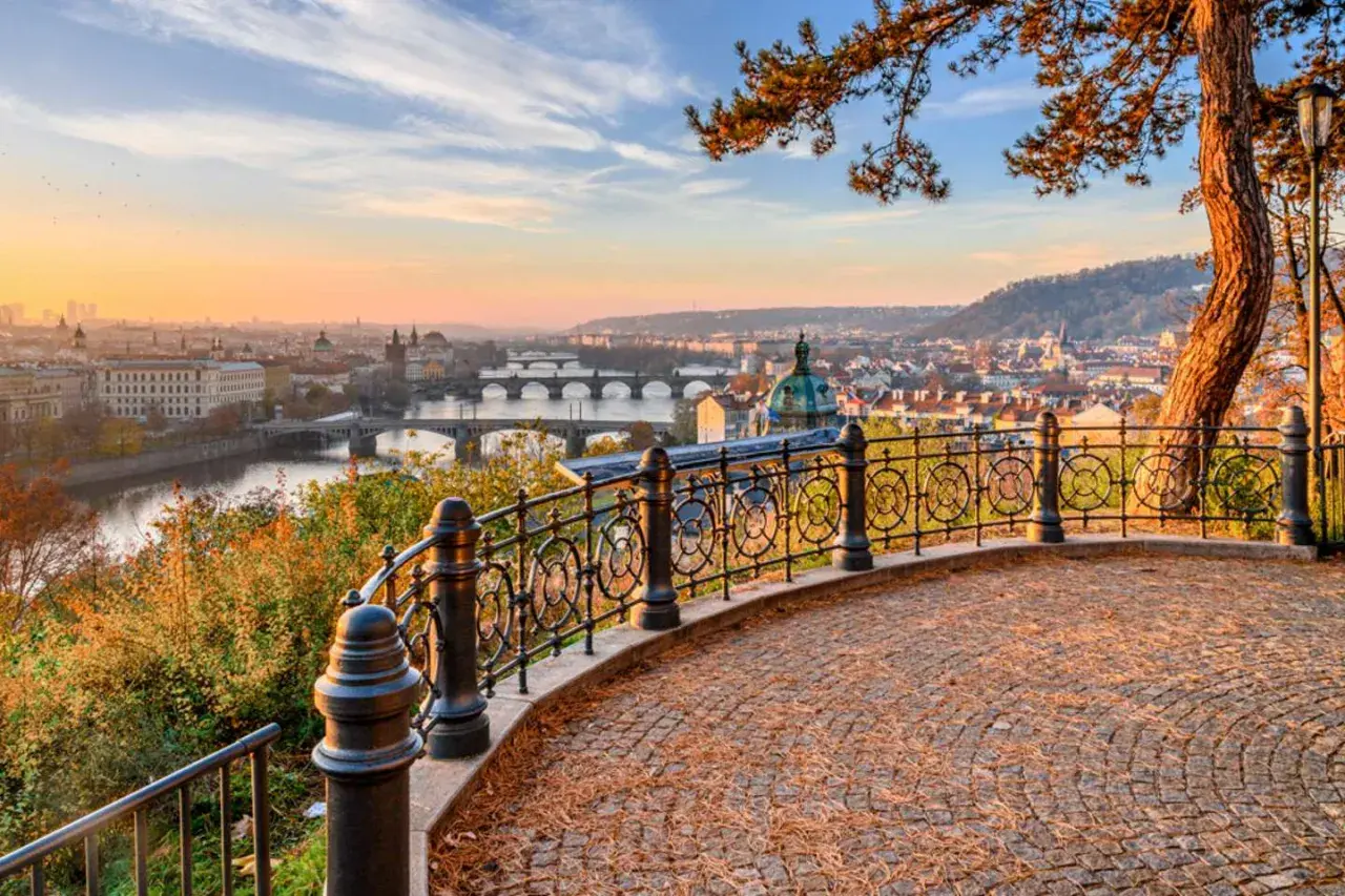 Prag im Herbst: Blick über die Moldau mit Brücken und Stadtkulisse bei Sonnenuntergang. Ein malerischer Ausblick von einem Aussichtspunkt mit gepflastertem Boden und schmiedeeisernem Geländer.