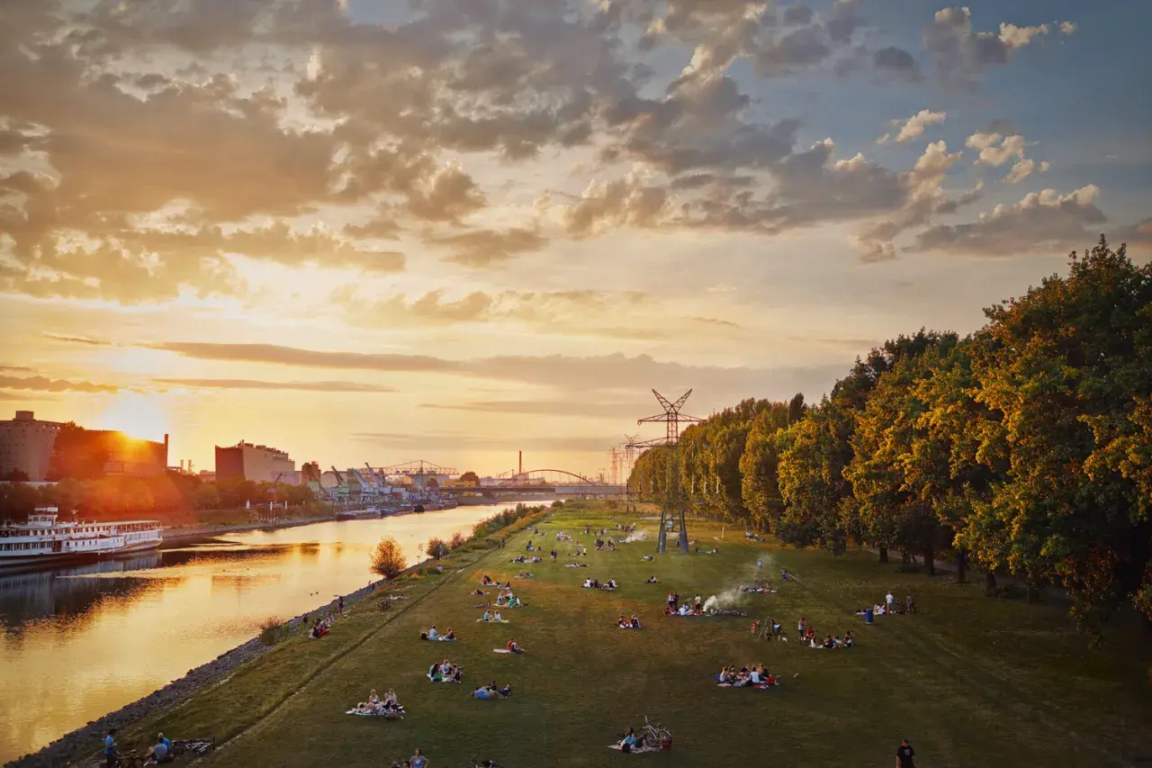 Sonnenuntergang an der Neckarwiese Mannheim: Menschen entspannen auf der Wiese, ein Schiff fährt auf dem Fluss, im Hintergrund Industrie und Bäume.
