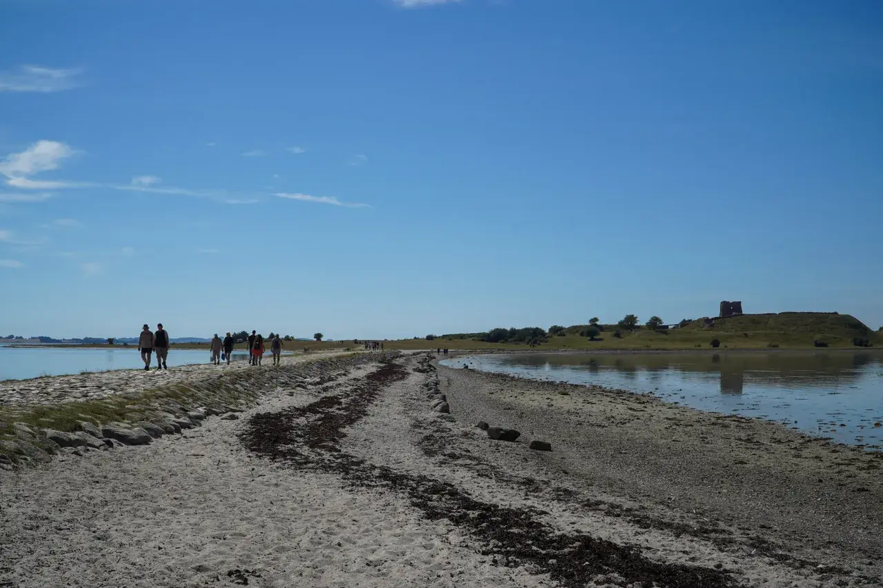 Menschen spazieren auf einem sandigen Weg entlang des Wassers in Richtung der Mols Bjerge mit einer alten Burgruine auf dem Hügel.