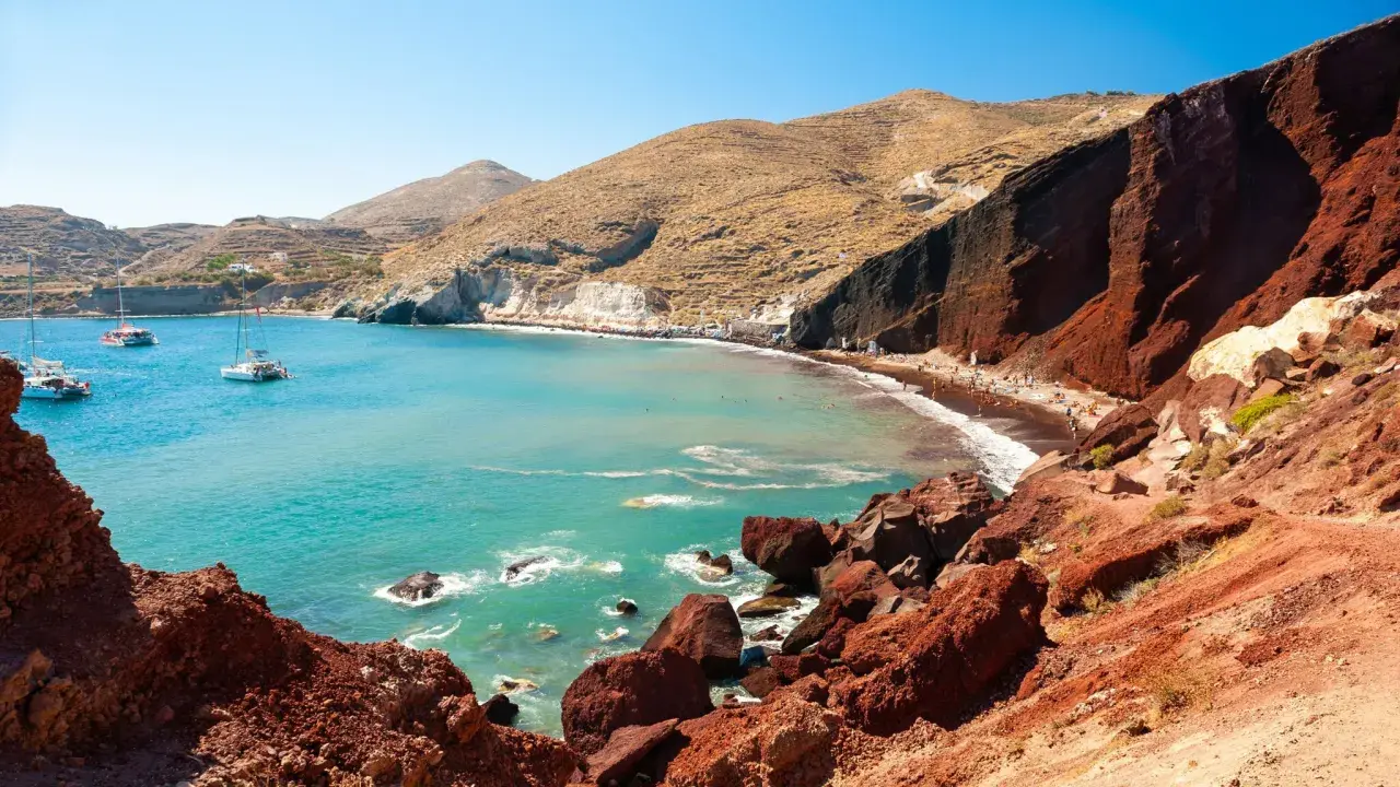 Der Rote Strand von Santorini mit türkisfarbenem Wasser, Segelbooten und Menschen, die sich auf dem roten Sand entspannen.