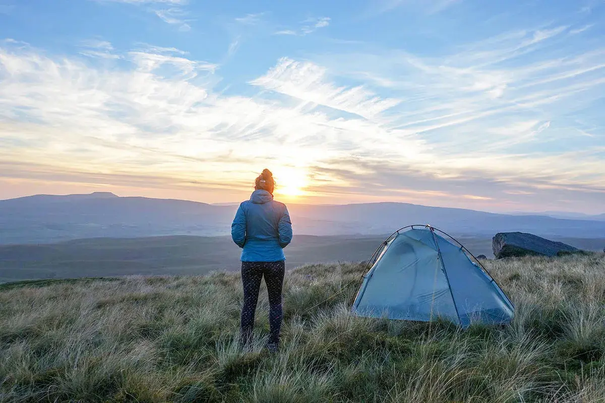 Frau blickt auf Sonnenuntergang, ihr Zelt steht bereit für das Wandern mit Zelt. Die Landschaft ist hügelig, der Himmel dramatisch.