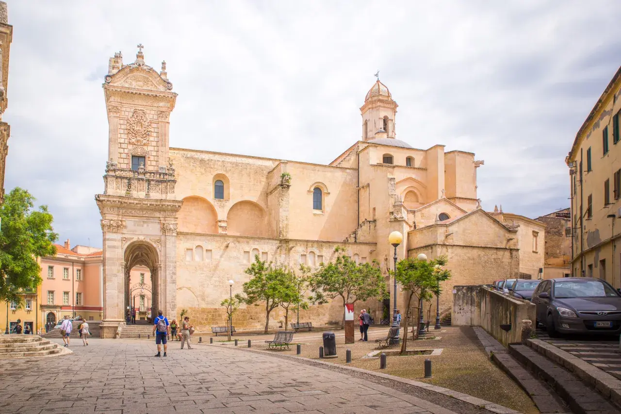 Ein sonniger Tag in der **Alghero Altstadt**. Menschen schlendern über den Platz vor einer imposanten Kirche mit einem verzierten Turm.
