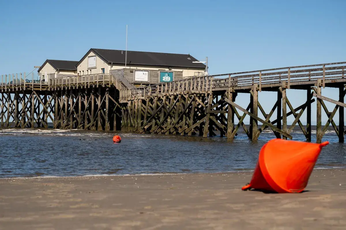 St. Peter-Ording an der Nordsee: Holzsteg mit Häusern, Bojen im Wasser und eine orangefarbene Boje am Strand.