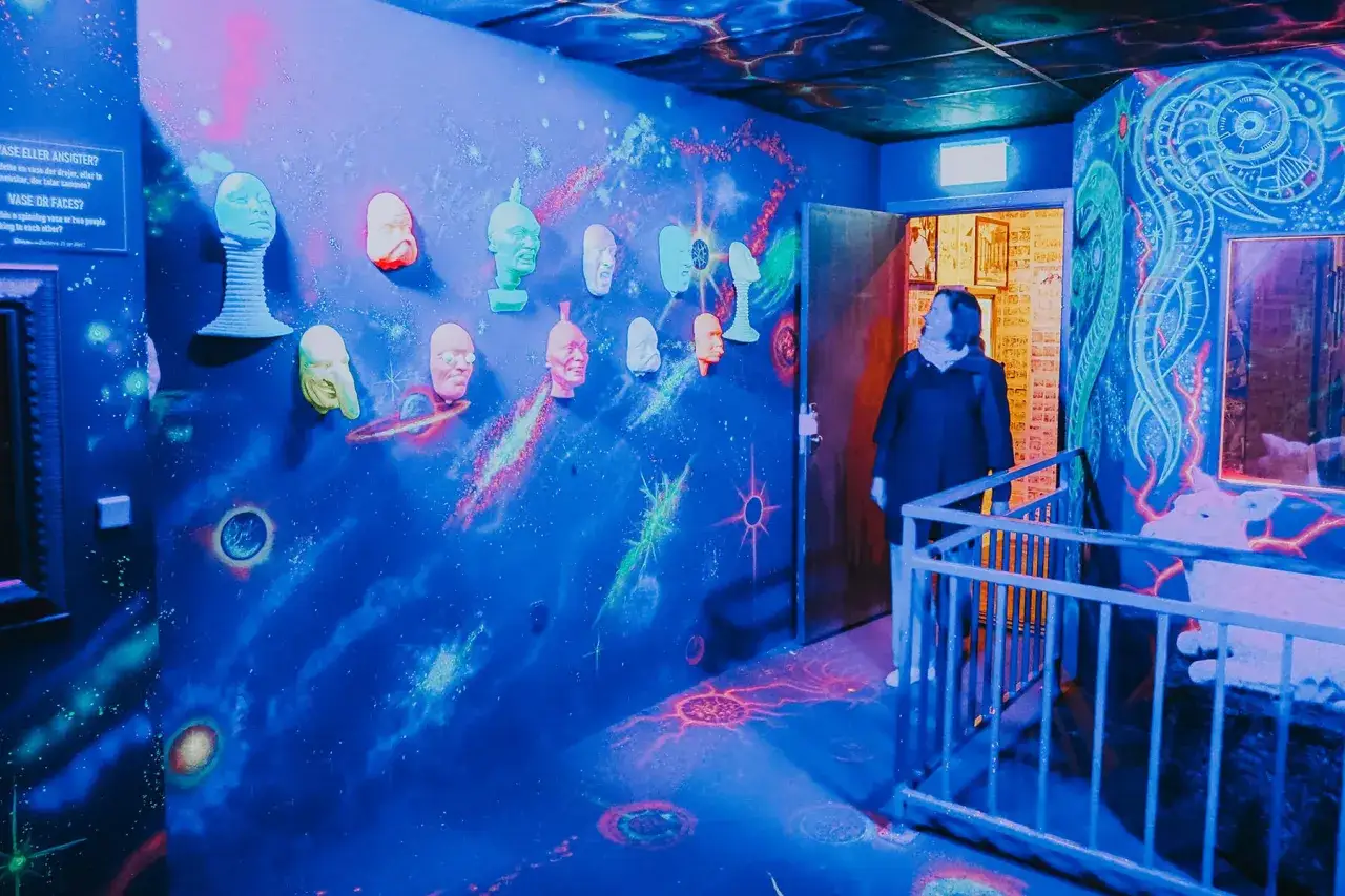 Kopenhagen bei Regen: Eine Frau steht in einem neonbeleuchteten Raum mit Weltraum-Motiven und vielen Gesichtern an der Wand.