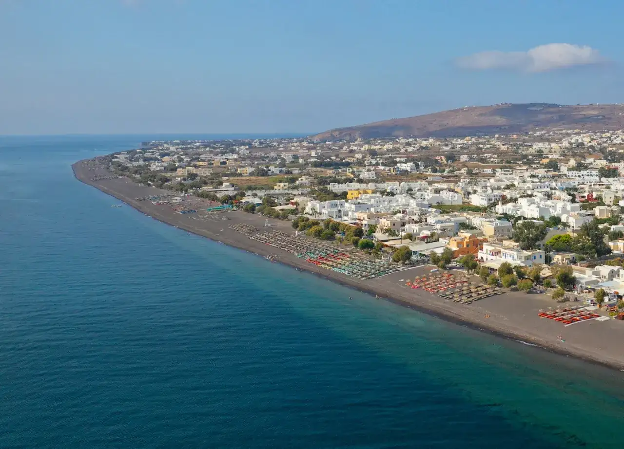 Blick auf den schwarzen Sandstrand von Santorini mit Reihen von Liegest&uuml;hlen und Sonnenschirmen. Wei&szlig;e H&auml;user s&auml;umen die K&uuml;ste, das tiefblaue Meer glitzert.