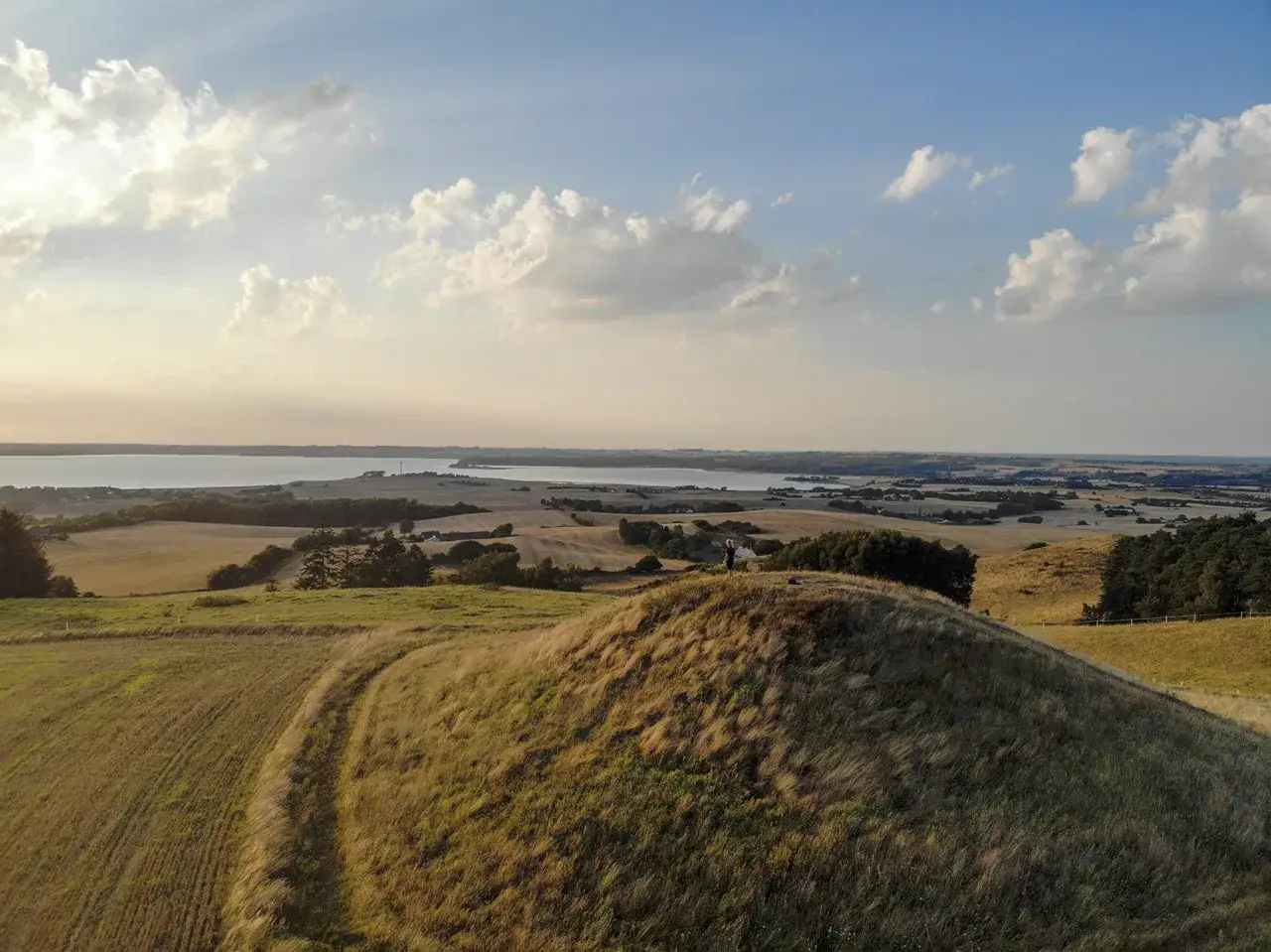 Blick über die sanften Hügel der Mols Bjerge bei Sonnenuntergang. Eine Person steht auf einem grasbewachsenen Hügel mit Blick auf das Meer und die umliegende Landschaft.