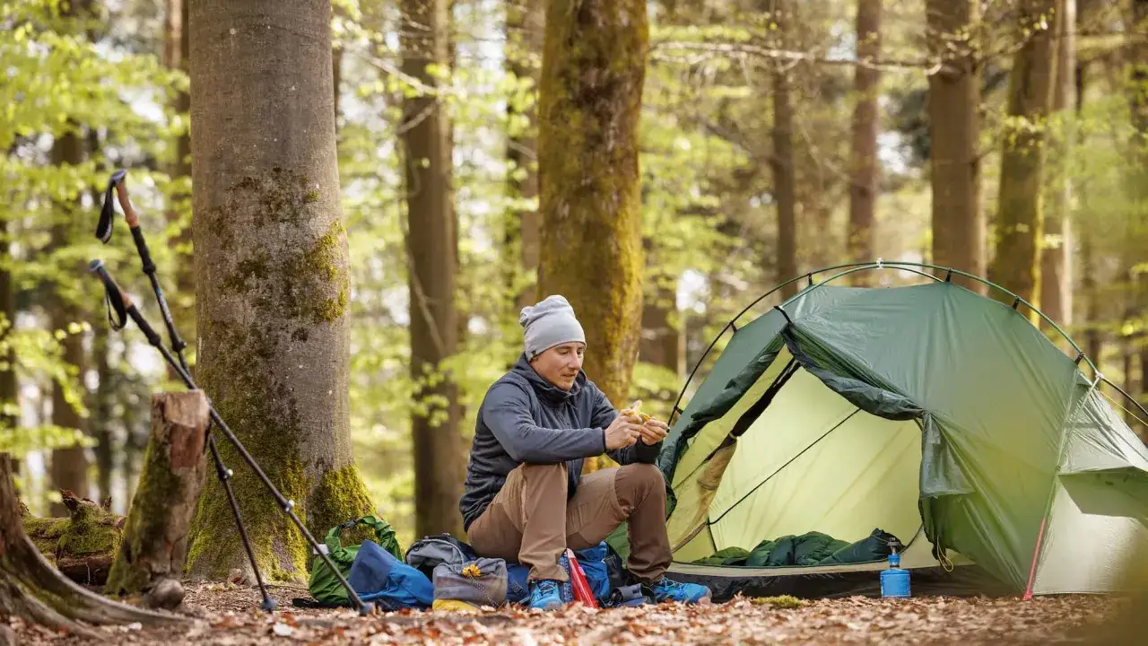 Mann sitzt neben gr&uuml;nem Zelt im Wald, isst einen Snack. Wanderst&ouml;cke und Rucks&auml;cke sind bereit f&uuml;r das n&auml;chste Abenteuer beim Wandern mit Zelt.