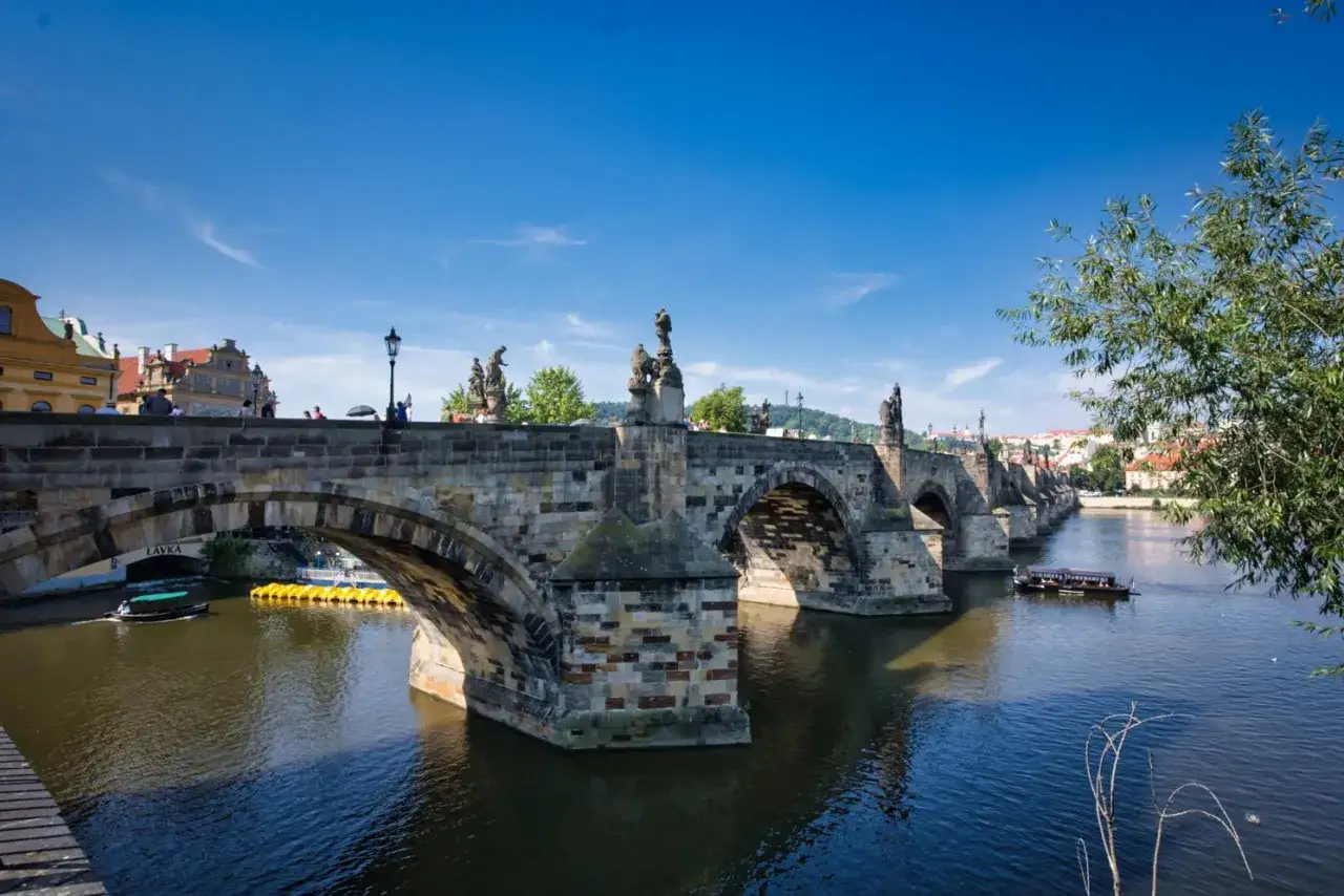 Karlsbr&uuml;cke in Prag im Sommer. Touristen spazieren &uuml;ber die Br&uuml;cke, w&auml;hrend ein Boot auf der Moldau f&auml;hrt.