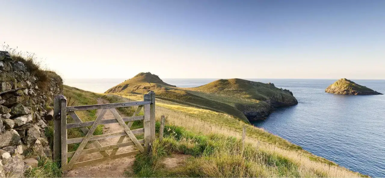 Ein Holztor öffnet den Weg entlang des South West Coast Path. Grüne Hügel und das Meer prägen die Landschaft.