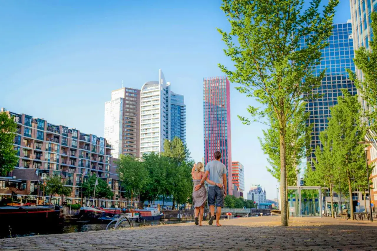 Ein Paar spaziert am Wasser entlang, vorbei an modernen Geb&auml;uden und Booten. Entdecken Sie die versteckten Sch&ouml;nheiten von Rotterdam!