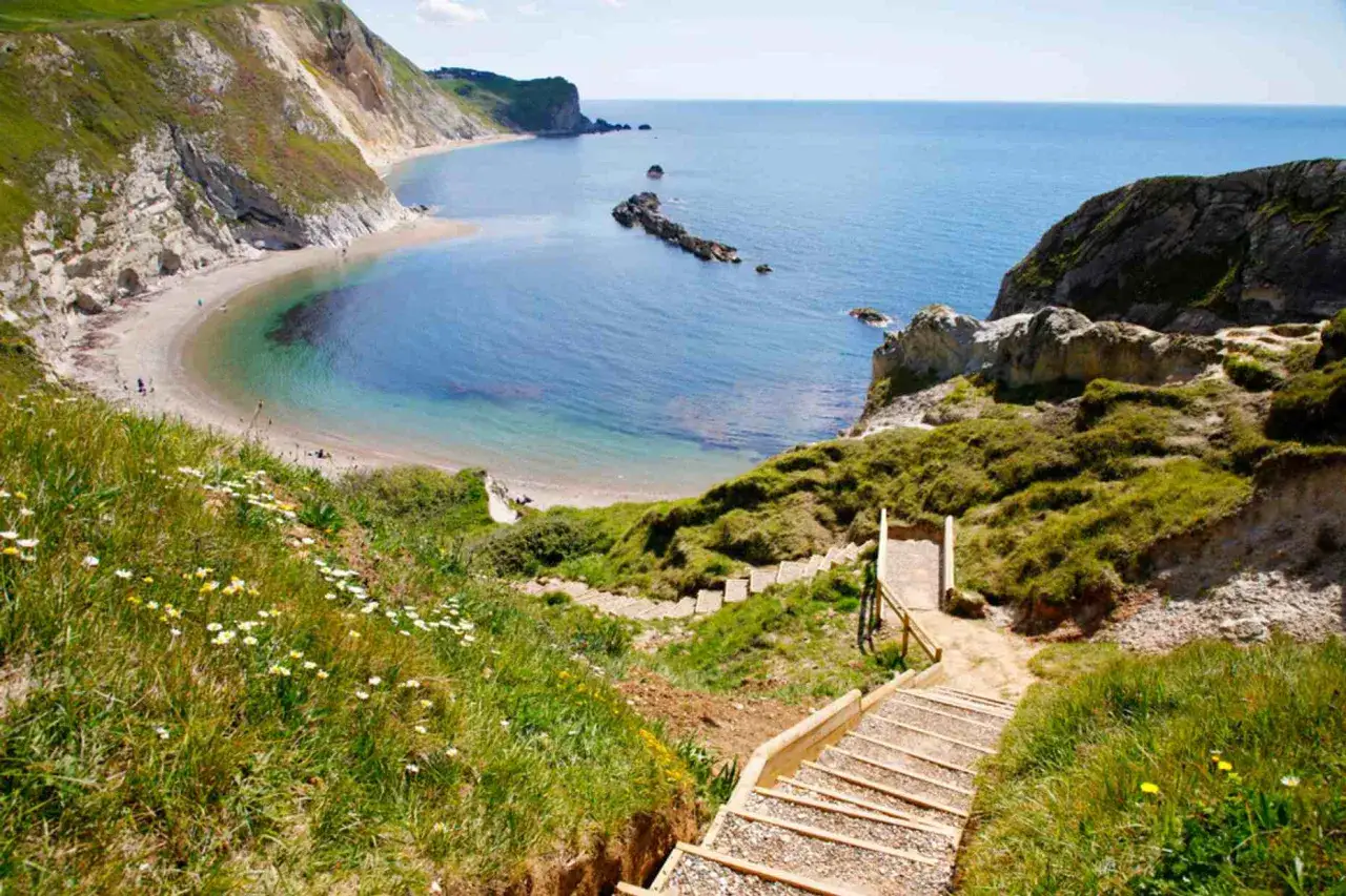 Treppen führen zu einer Bucht am South West Coast Path. Grüne Hänge mit Blumen und das blaue Meer.