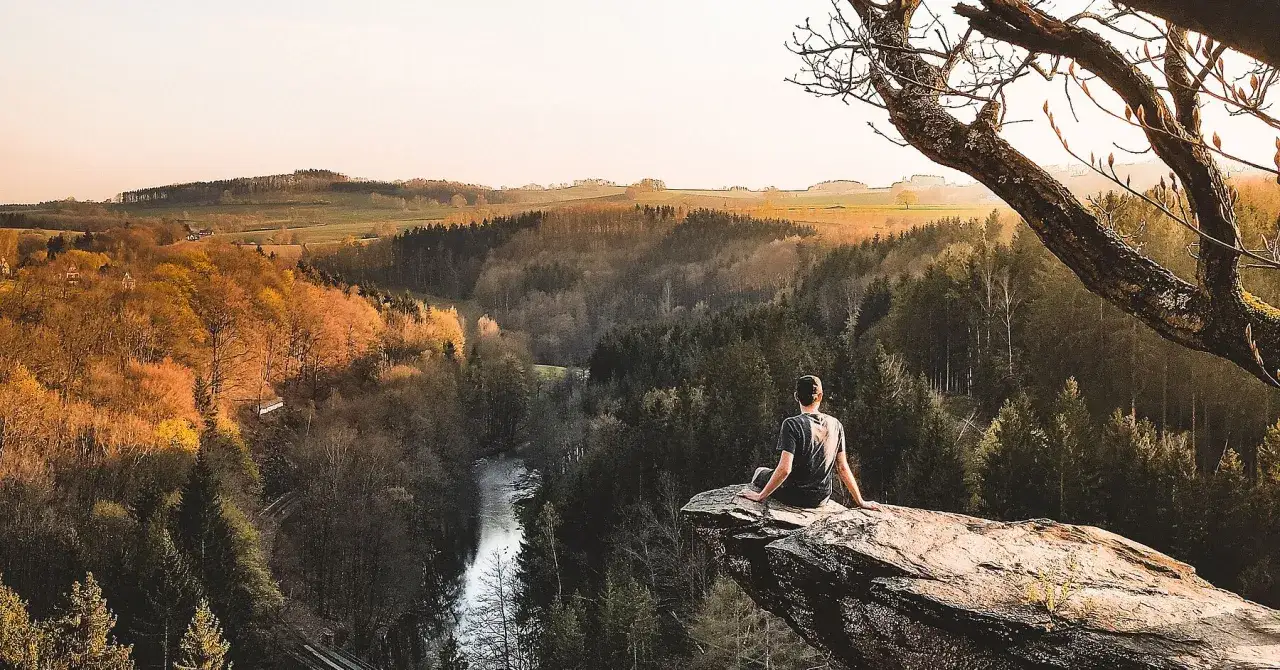 Ein Mann sitzt auf einem Felsvorsprung und blickt ins Tal. Ein Fluss schlängelt sich durch den Wald. Ein echter Geheimtipp im Erzgebirge für Naturliebhaber.