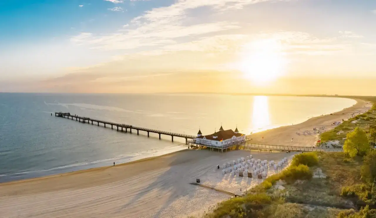 Sonnenuntergang an der Ostsee in Deutschland. Ein langer Steg führt zu einem weißen Gebäude am Strand, daneben Liegestühle.