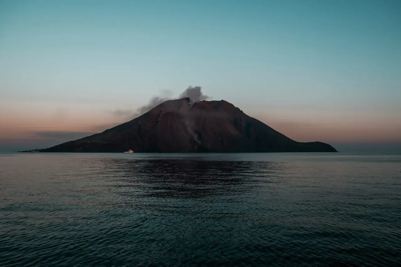 Rauchender Vulkan auf einer Insel im Meer, ein Schiff n&auml;hert sich. Ein Beispiel f&uuml;r die beeindruckenden Vulkanlandschaften Europas.