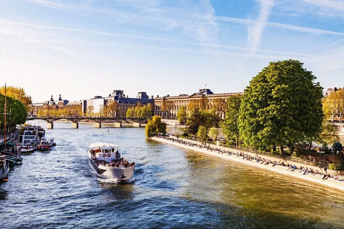 Ein sonniger Tag in Paris im Sommer. Ein Ausflugsboot f&auml;hrt auf der Seine, im Hintergrund das Louvre-Museum und Menschen, die am Ufer entspannen.
